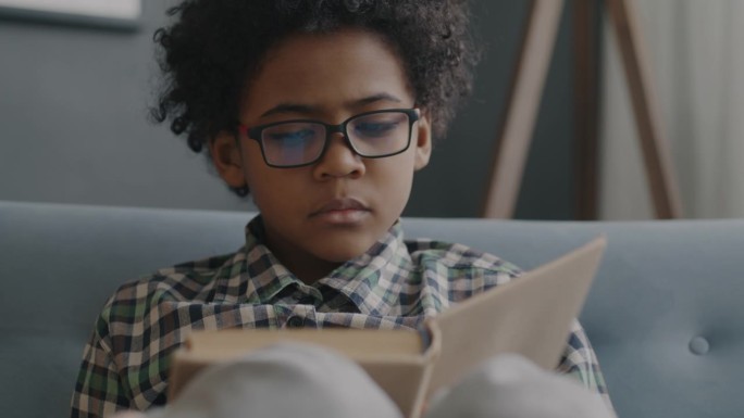 聪明的非裔美国男孩戴着眼镜在家里看书微笑的慢镜头