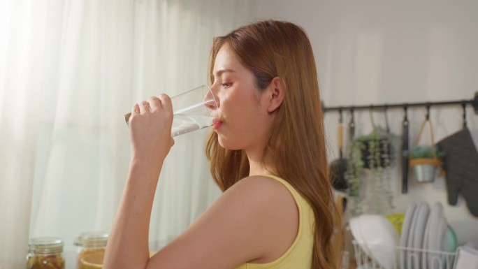 亚洲年轻漂亮的女性在家里的厨房喝一杯水。年轻口渴的美丽女孩在早上醒来后手握干净的矿泉水杯，轻啜一口以
