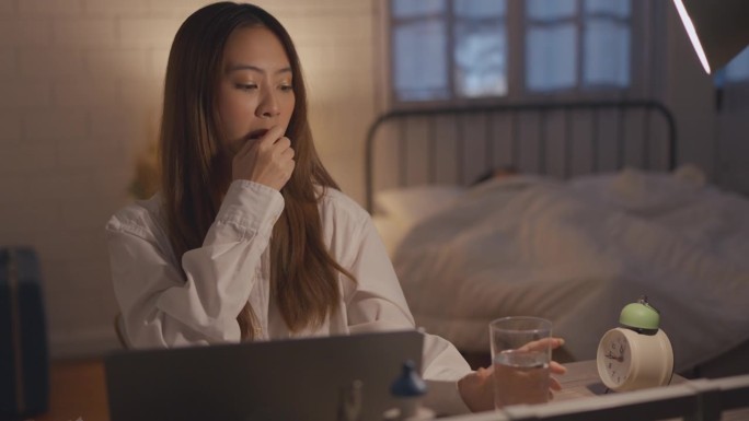 疲惫不堪的亚洲年轻女子工作过度，晚上还要吃药。迷人的女商人在家里的卧室里用笔记本电脑上网时感到恶心和
