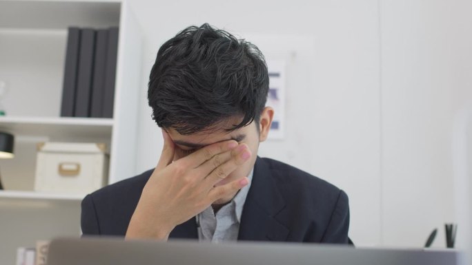 亚洲商人在现代办公室里使用笔记本电脑工作时，穿着西装，疲惫不堪