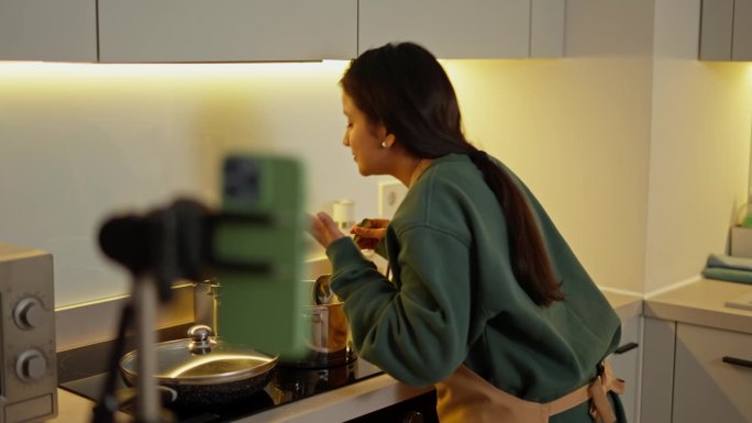 一个快乐的黑发女孩，棕色皮肤，穿着绿色夹克，系着棕色围裙，一边在现代厨房里的一个灰色闪亮的平底锅里做