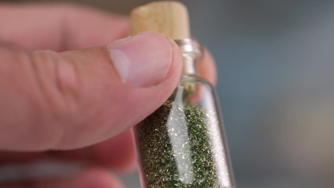男性的手指拿着一个装着闪光剂的小瓶，放在一个玻璃容器里，转动小瓶，以慢动作观察闪闪发光的微粒。
