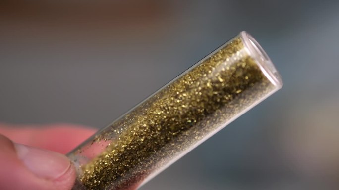 一个男人的手指小心地转动着一个玻璃小瓶，在一个微距的慢动作镜头中捕捉到闪闪发光的颗粒。