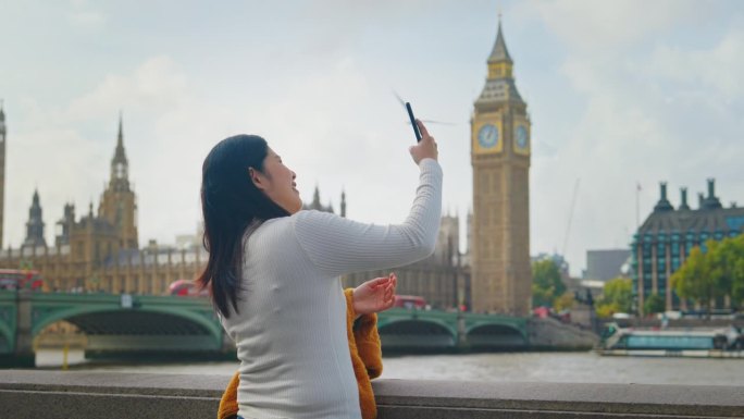 在英国伦敦，一名亚洲女游客用手机站在泰晤士河边，以大本钟和国会大厦为背景，开心地拍照