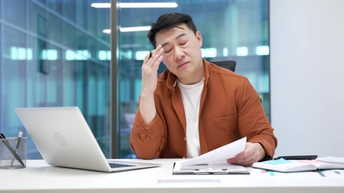 疲惫不堪的亚洲商人因在现代商务办公室的桌子上处理文书工作而感到沮丧。