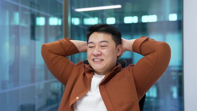 微笑的亚洲男性专业人士在办公室放松，靠在椅子上。