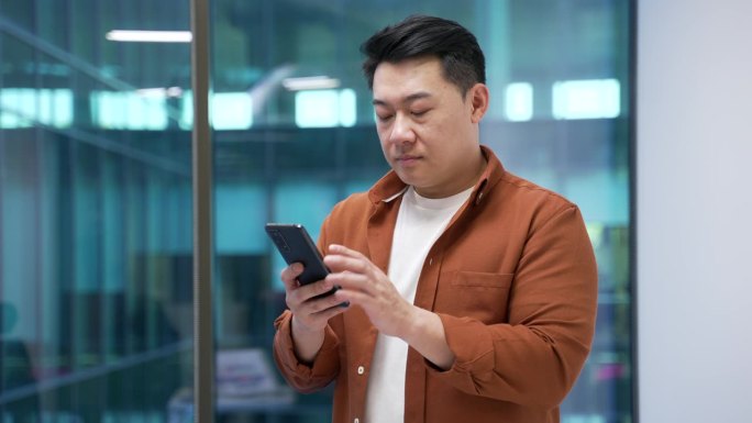 亚洲商人在现代商务办公室内站着使用手机浏览。