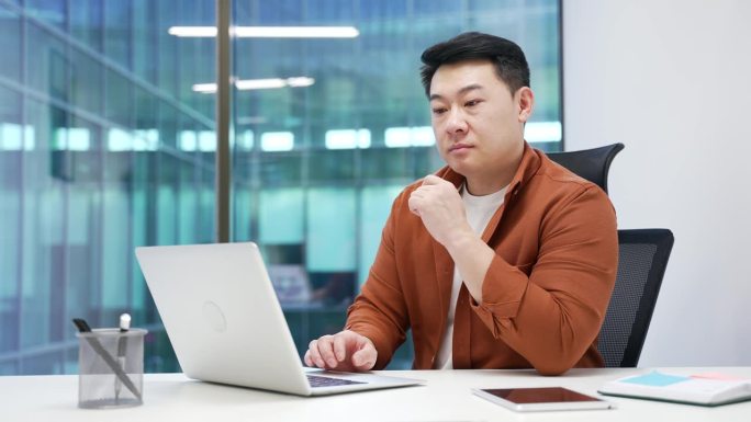专注的亚洲商人在现代办公室分析工作，沉思的专业人士，坐在桌子前用电脑打字。