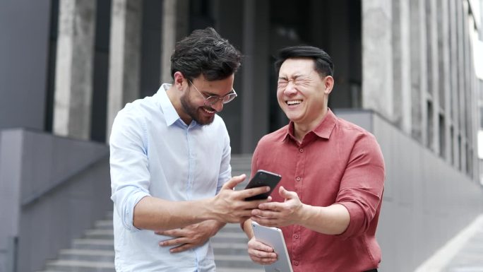 两位商人在办公楼外看手机，享受幽默时刻。