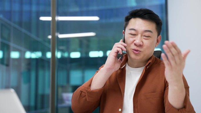 自信的亚洲商人在现代办公室的工作场所坐着，打着手机。