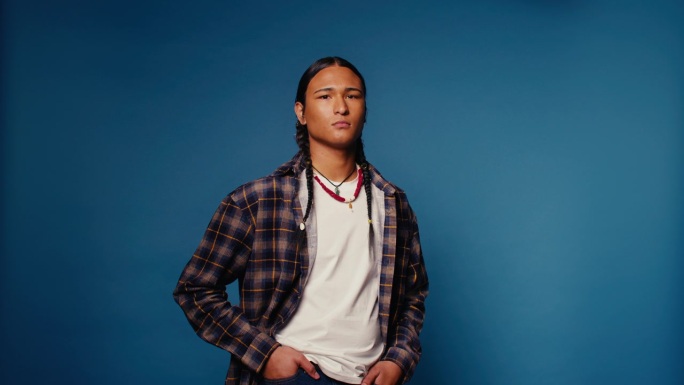 年轻男子扎着辫子，佩戴项链，在蓝色背景的摄影棚中严肃地摆姿势。