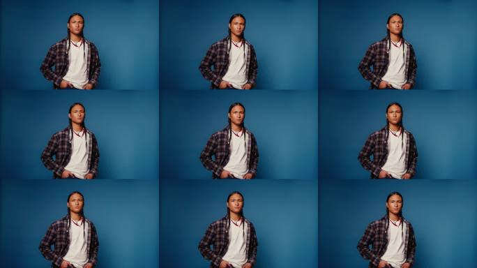 年轻男子扎着辫子，佩戴项链，在蓝色背景的摄影棚中严肃地摆姿势。