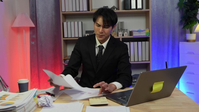 一名西装革履的亚裔商务人士因工作未按计划完成而沮丧地将一份揉皱的文件扔掉。