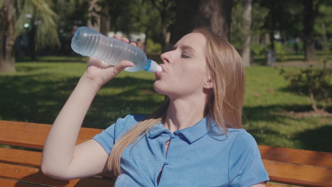 年轻女性在公园里与炎热作斗争，通过喝水和将水瓶涂抹在脸上来降温。她坐在阴凉的长椅上，寻求夏季的凉爽。