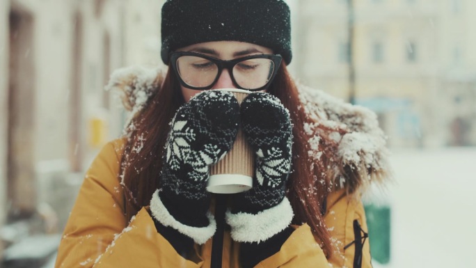 特写镜头：一位美丽的棕发女孩戴着眼镜，在冬季城市中漫步，喝着热咖啡。一位微笑的女性在冬季城市街道上走