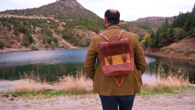 一位戴眼镜、背着皮质背包的男士面带微笑，走向乡村中平静的湖面，镜头以4K分辨率平滑地围绕他旋转。
