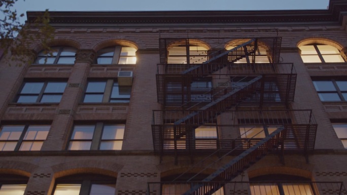 风化的砖砌外立面与金属消防梯在黄昏街景中投射出戏剧性的阴影，捕捉到典型的纽约建筑叙事。