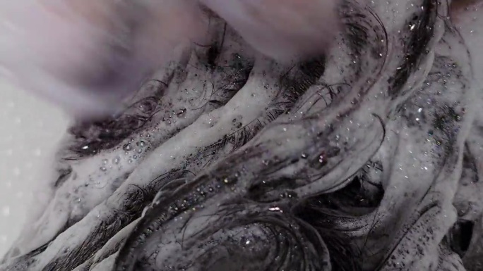 女人纤长乌黑的秀发，伴随丰富泡沫的洗发水，特写镜头。护发和清洁过程的视频。女性双手按摩头皮和头发，产