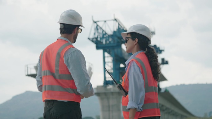 身穿安全背心和头盔的土木工程师在桥梁施工现场使用测绘望远镜设备，讨论基础设施发展的项目计划。
